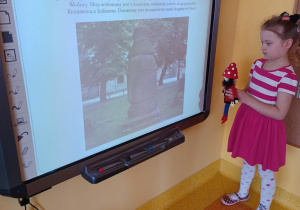 Dziewczynka stoi z maskotką Krasnala Hałabały pod tablicą interaktywną na której wyświetlone jest zdjęcie Słupa Milowego.